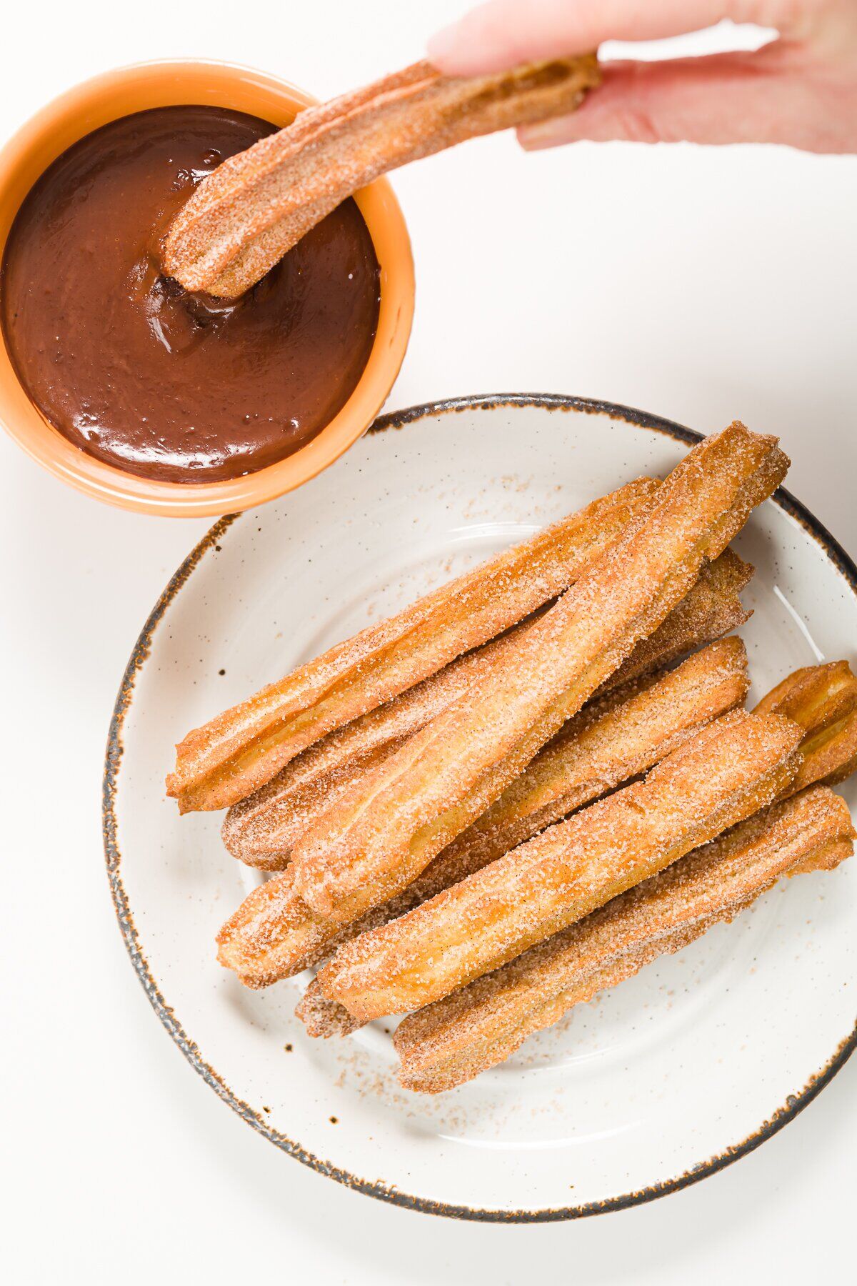 Overhead shot of air fryer churros on a white plate and one being dipped into a bowl of chocolate sauce