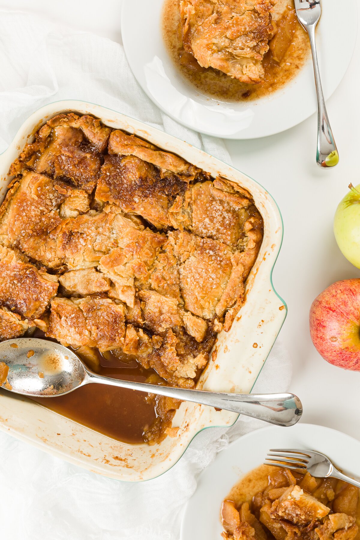 Overhead shot of pan of apple pandowdy, plates with food, and apples