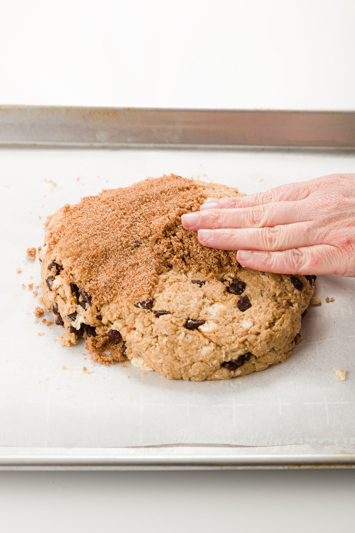 Circle of scone dough on a parchment-lined cookie sheet with hand spreading cinnamon sugar mixture on top of it