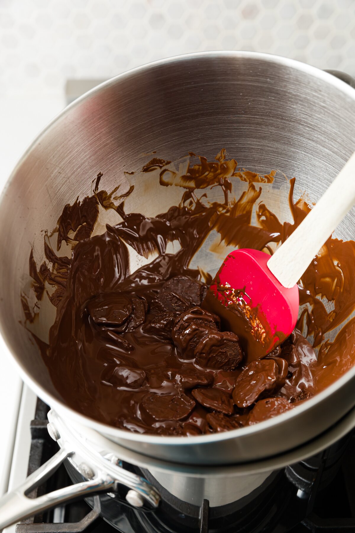 partially melted chocolate in a mixing bowl over a pot on the stove with a red spatula
