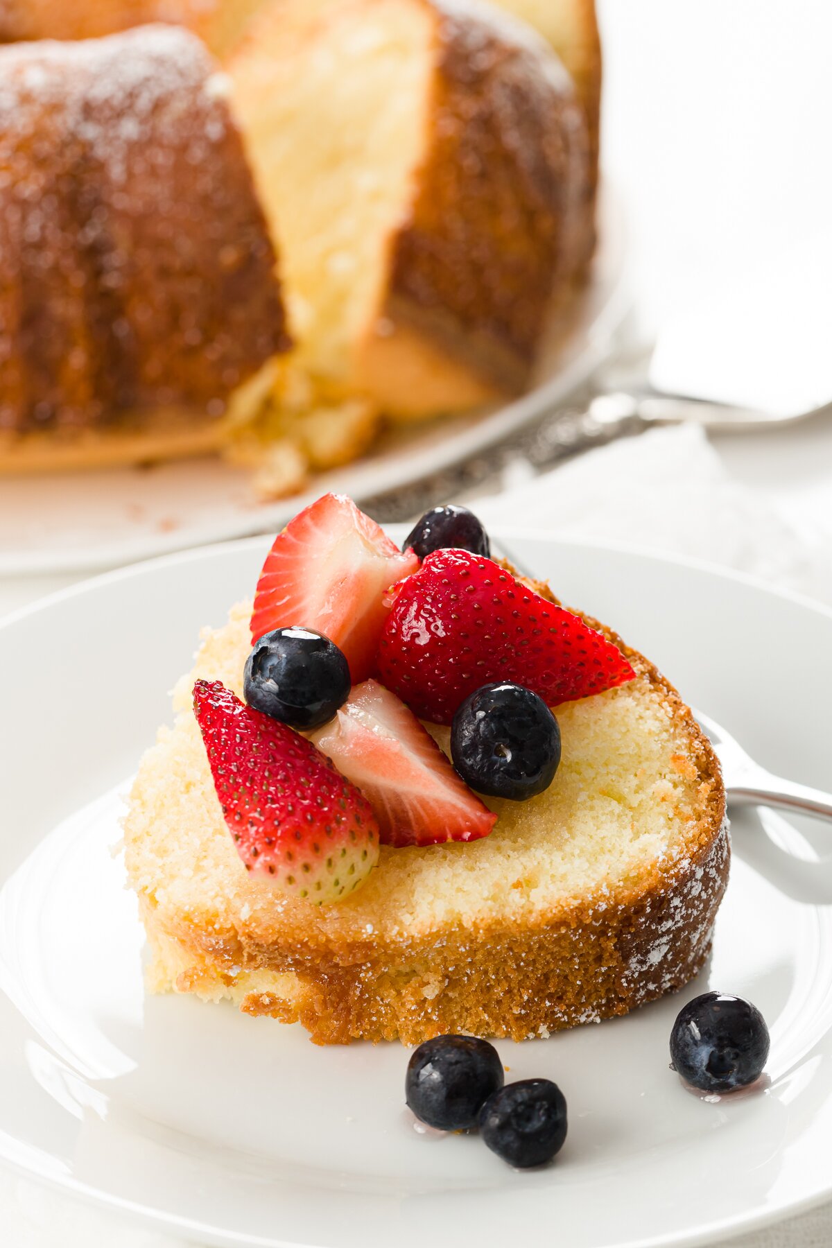 a slice of pound cake covered in berries with the full cake in the background